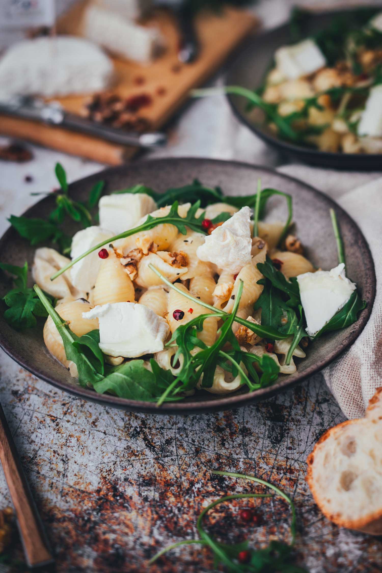 Seitliche Nahaufnahme von einem Teller, gefüllt mit cremiger Pasta mit Ziegenkäse, garniert mit Rucola und Walnüssen. Im Vordergrund sieht man etwas Baguette, im HIntergrund einen weiteren Teller sowie ein Holzbrett mit Ziegenkäse.