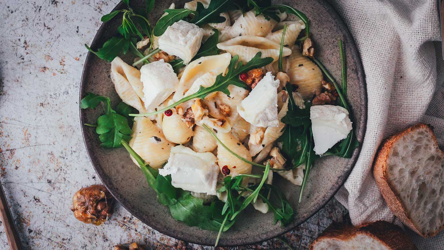 Topshot Nahaufnahme von einem Teller mit Muschelnudeln in einer Ziegenkäse-Sauce, serviert mit frischem Rucola und karamellisierten Walnüssen. Daneben liegt frisches Brot zum Servieren.