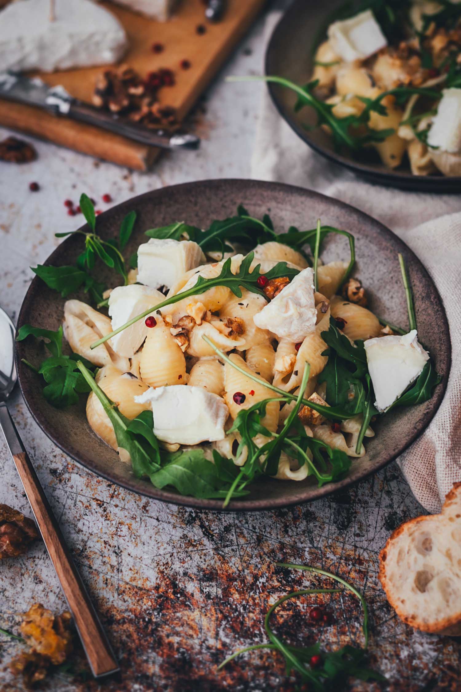 Pasta mit Ziegenkäse auf einem Teller, serviert und in seitlicher Ansicht. Man sieht wunderbar cremige Muschelnudeln mit dem hellen Ziegenkäse, dazu knackigen Rucola und knusprige Walnusskerne. Im Hintergrund ist ein zweiter Teller sowie ein Brettchen mit Ziegenkäse, im Vordergrund sieht man Nüsse und Baguettescheiben.