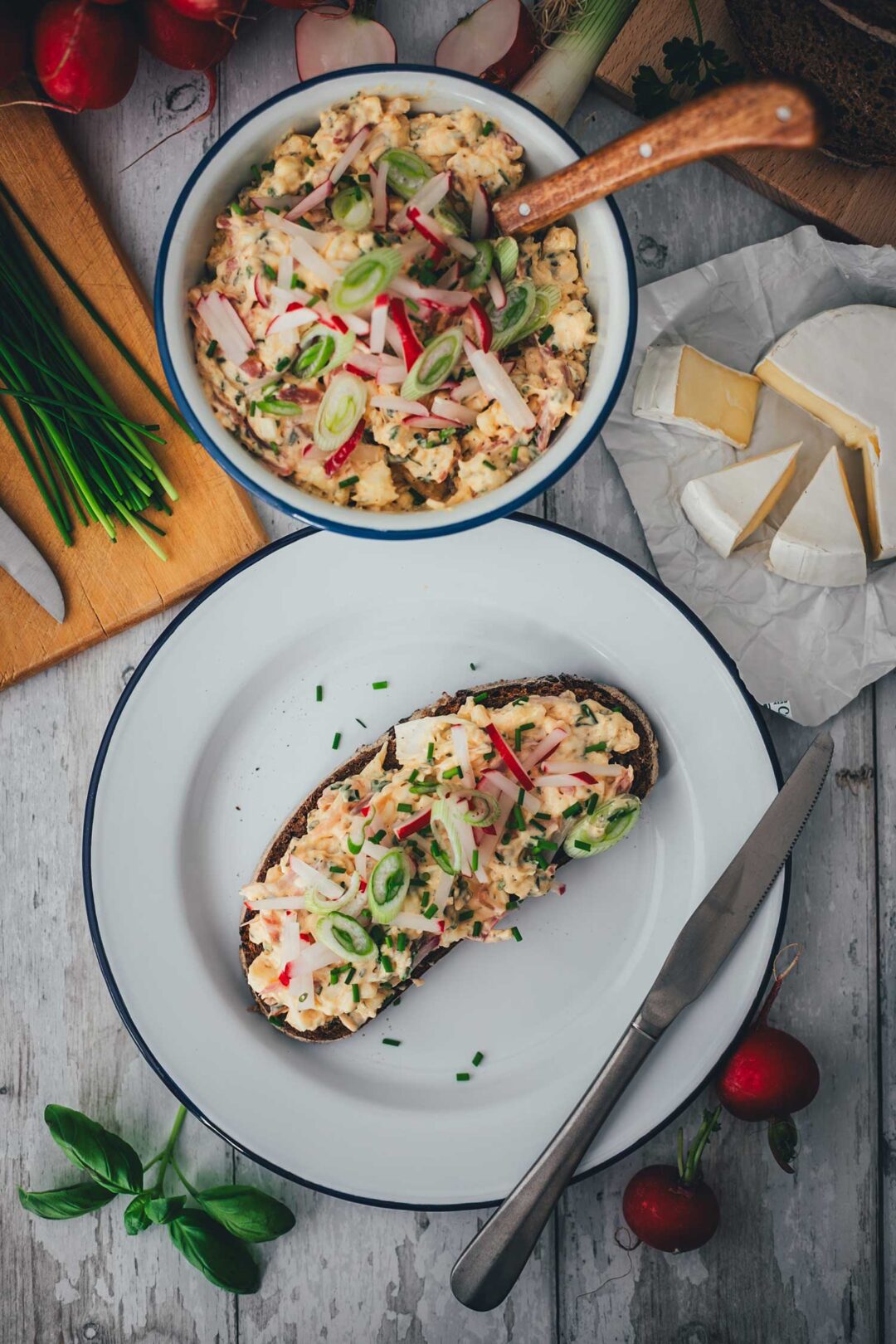 Draufsicht auf eine gemütliche Frühlings-Brotzeit: Eine reich belegte Scheibe Sauerteigbrot mit Obatzda, eine Schüssel mit dem Aufstrich, frische Camembert-Ecken, Radieschen und Kräuter auf einem hellen Holztisch.