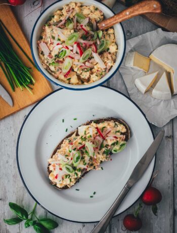 Draufsicht auf eine gemütliche Frühlings-Brotzeit: Eine reich belegte Scheibe Sauerteigbrot mit Obatzda, eine Schüssel mit dem Aufstrich, frische Camembert-Ecken, Radieschen und Kräuter auf einem hellen Holztisch.
