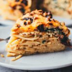 Seitliche Nahaufnahme von einem Stück Spaghetti-Kuchen. Nudelauflauf wird in einer Springform in Kuchenform gebacken und anschließend in Form von Kuchenstücken serviert. Man sieht die einzelnen Schichten von Spaghetti, Spinat und getrockneten Tomaten. Im Hintergrund ist der ganze Spaghettikuchen zu sehen.