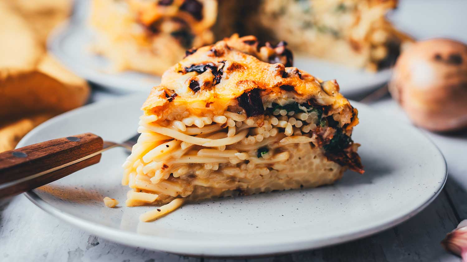 Seitliche Nahaufnahme von einem Stück Spaghetti-Kuchen. Nudelauflauf wird in einer Springform in Kuchenform gebacken und anschließend in Form von Kuchenstücken serviert. Man sieht die einzelnen Schichten von Spaghetti, Spinat und getrockneten Tomaten. Im Hintergrund ist der ganze Spaghettikuchen zu sehen.