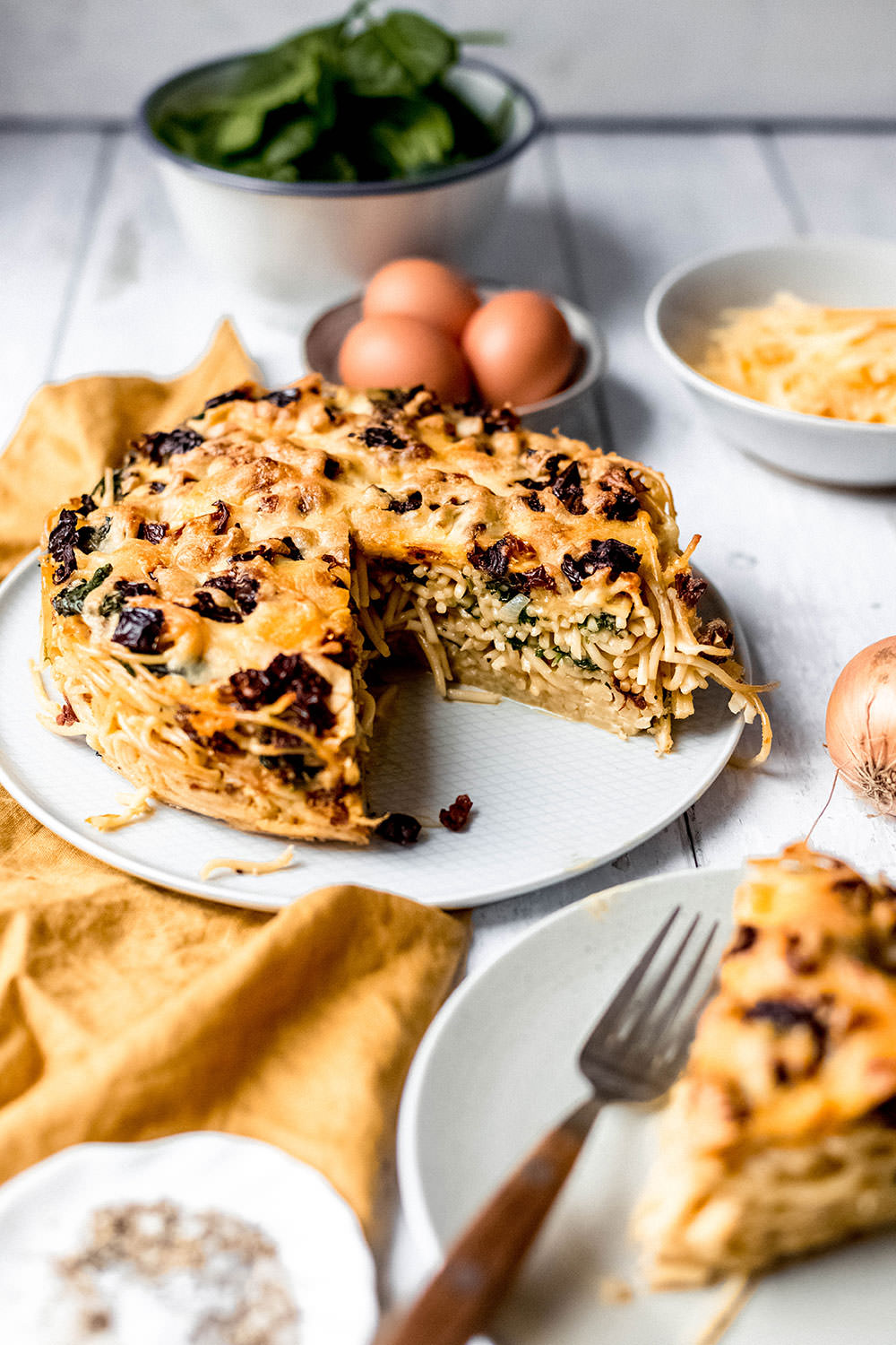 Nahaufnahme auf den runden Spaghetti-Kuchen aus der Springform. Es wurde ein Stück entnommen und man guckt direkt auf den Anschnitt, sieht die Spaghetti, den Spinat und die getrockneten Tomaten.