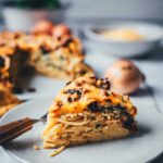 Auf einem Teller liegt ein Stück Spaghetti-Kuchen. Das ist ein Nudelauflauf aus Spaghetti, Spinat und getrockneten Tomaten, der in einer Springform gebacken und wie in ein Stück Kuchen geschnitten serviert wird. Man sieht die cremigen Schichten aus allen Zutaten und den ganzen Kuchen im Hintergrund.