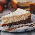 Nahaufnahme von einem Stück des Carrot Cake Cheesecakes auf einem marmomierten Teller. Man sieht sehr gut die Schichten aus Rüblikuchen als Boden und dem typischen Cheesecake-Topping. Im HIntergrund steht der angeschnittene Kuchen, daneben liegen ein paar Möhren.