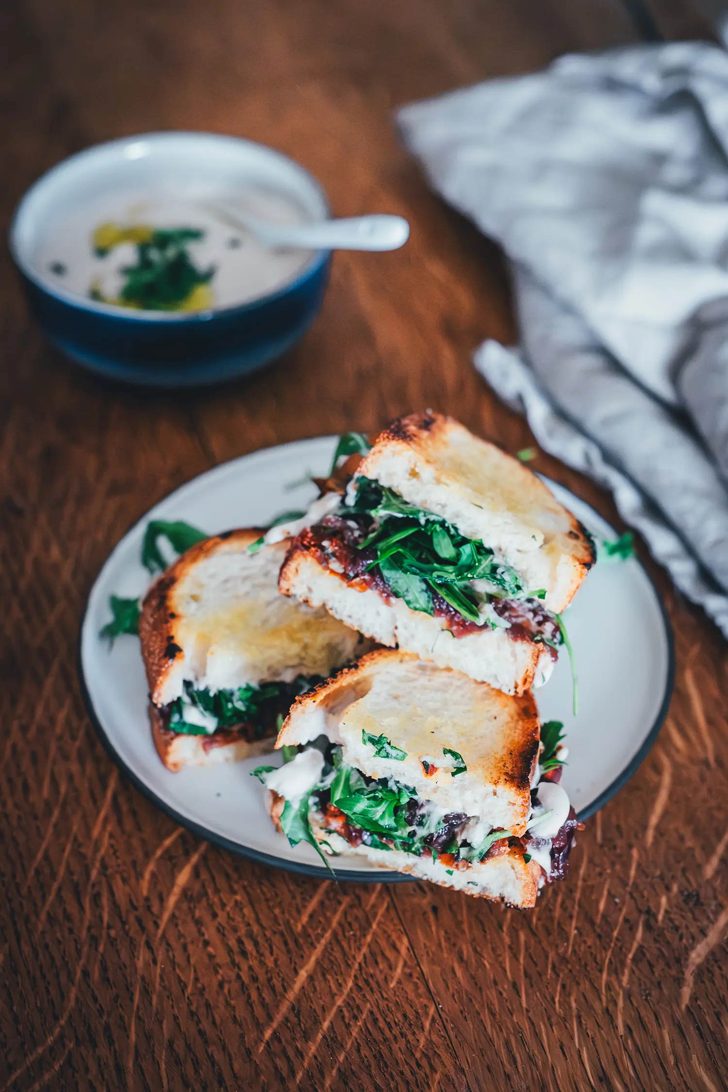 Drei belegte Hummus-Sandwiches liegen auf einem hellen Teller, im Hintergrund liegt eine Serviette und eine Schale mit weiterem Hummus.