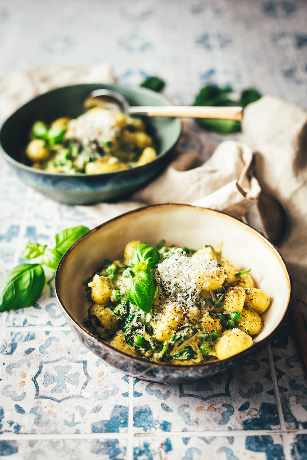 Ein Teller mit cremigen Gnocchi, mit Spinat und Erbsen, mit Pesto und geriebenem Käse. Serviert mit frischem Basilikum. Im Hintergrund steht eine weitere Portion.