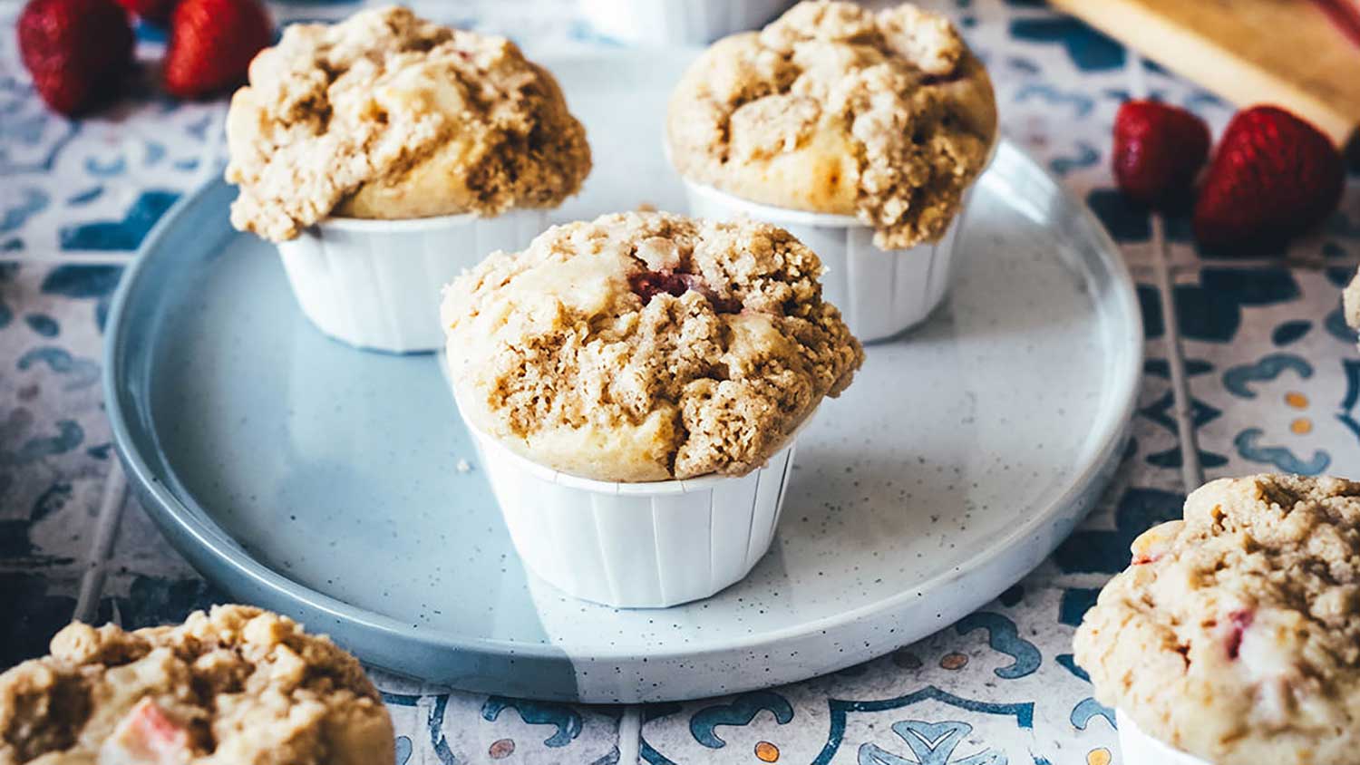 Frisch gebackene Erdbeer-Rhabarber-Muffins mit knusprigen Butterstreuseln stehen auf einem Teller. Im Hintergrund liegen frische Erdbeeren und Rhabarber.