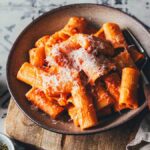 Eine Portion cremige Pasta alle Vodka auf einem Teller angerichtet. Sahnige Tomatensauce aus dem Vorrat mit Käse bestreut auf einem Küchenfliesen-Hintergrund mit einer grauen Serviette angerichtet.