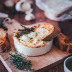 Nahaufnahme von einer Holzschachtel mit Camembert, der im Airfryer cremig zerlaufen ist. Getoppt mit Honig und Thymian und einem knusprigen Brot, das in den zerfließenden Käse getunkt wird. Im Hintergrund sieht man frischen Thymian, Knoblauch und weiteren Käse.