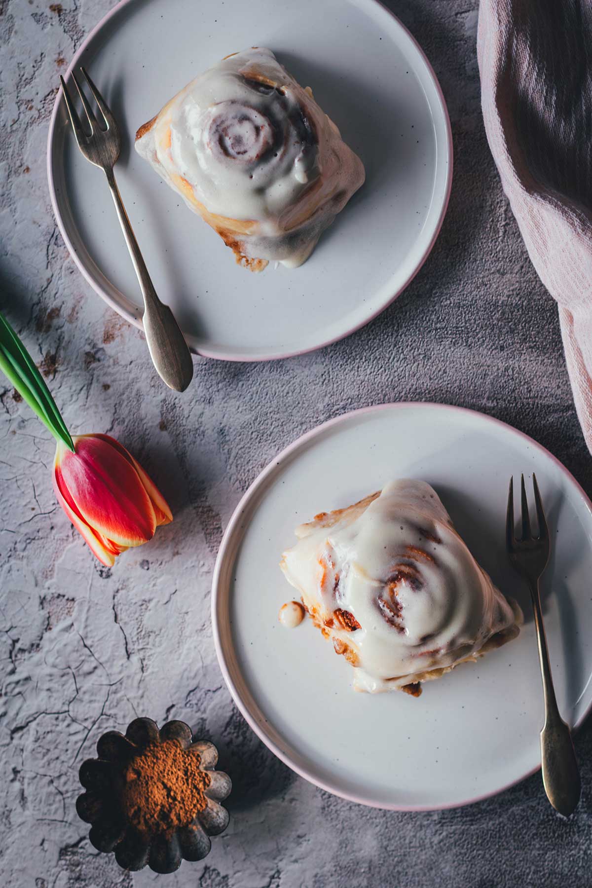 Top-Shot Aufnahme von zwei einzelnen Soft Cinnamon Rolls, die auf hellen Teller serviert werden. Daneben liegen eine blühende Tulpe und eine rosafarbene Serviette.