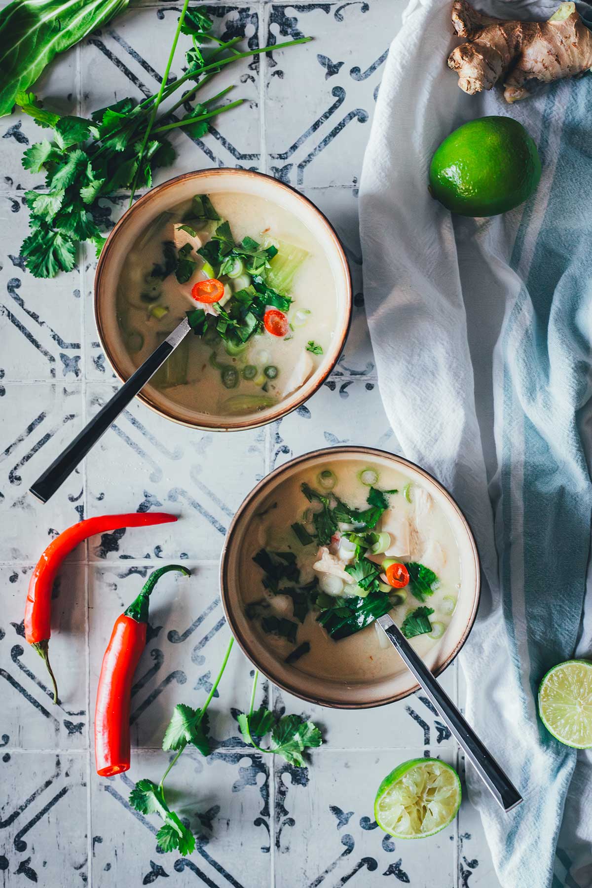 Zwei Schalen Thai-Kokos-Suppe mit Huhn, Pak Choi und Chili, serviert mit Limetten, Ingwer und Koriander auf einem rustikalen Untergrund | moeyskitchen.com