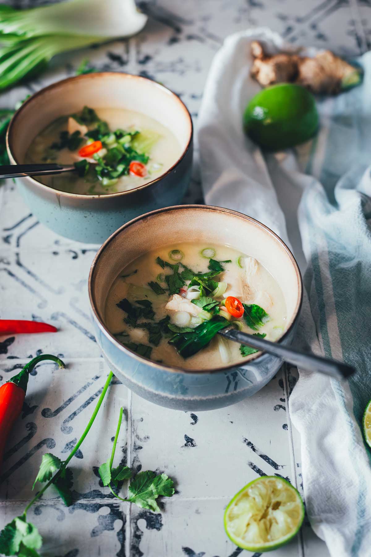 Zwei Schalen Thai-Kokos-Suppe mit Huhn, Pak Choi und Chili, serviert mit Limetten, Ingwer und Koriander auf einem rustikalen Untergrund | moeyskitchen.com