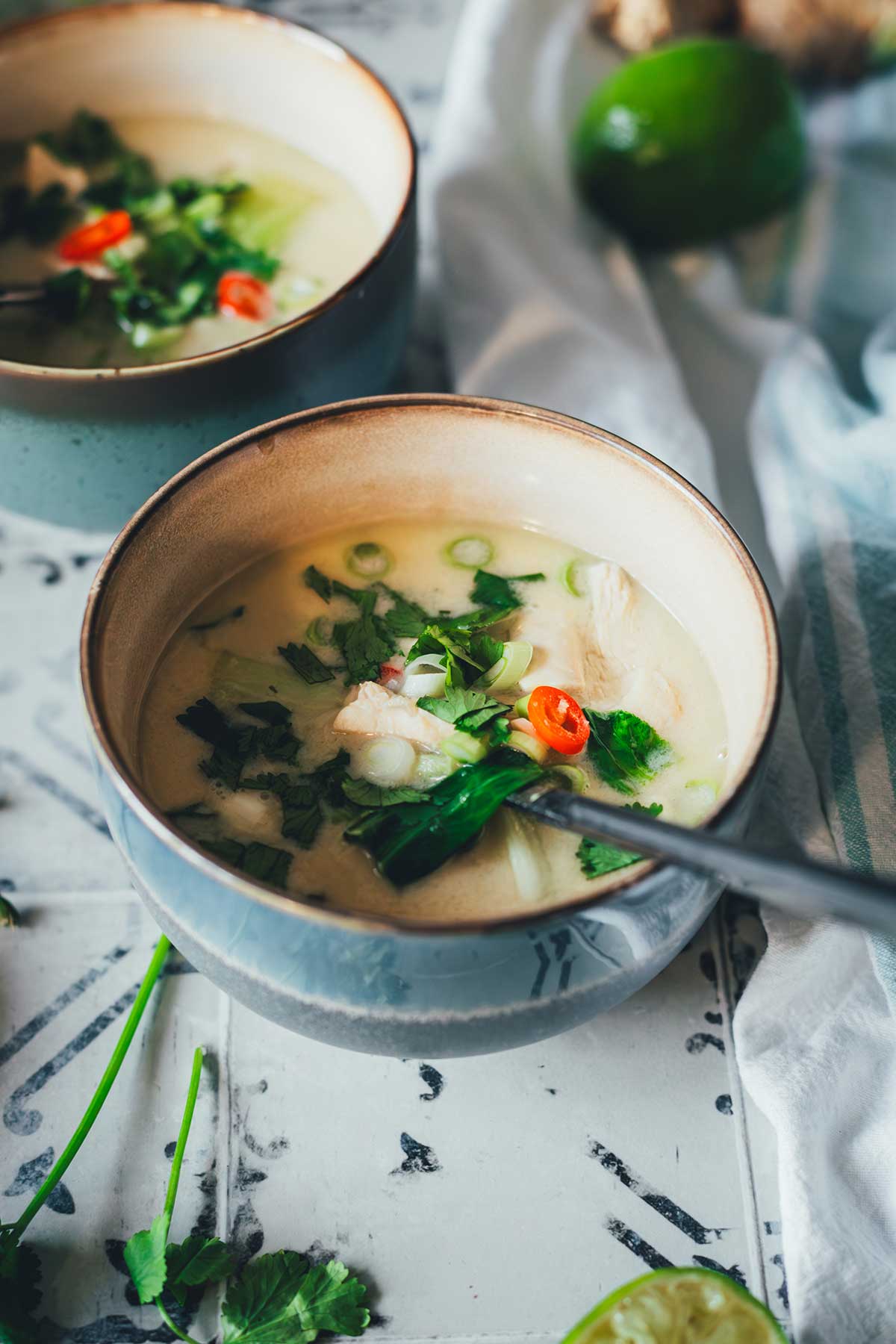 Zwei Schalen Thai-Kokos-Suppe mit Huhn, Pak Choi und Chili, serviert mit Limetten, Ingwer und Koriander auf einem rustikalen Untergrund | moeyskitchen.com