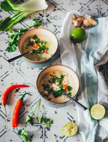 Zwei Schalen Thai-Kokos-Suppe mit Huhn, Pak Choi und Chili, serviert mit Limetten, Ingwer und Koriander auf einem rustikalen Untergrund | moeyskitchen.com