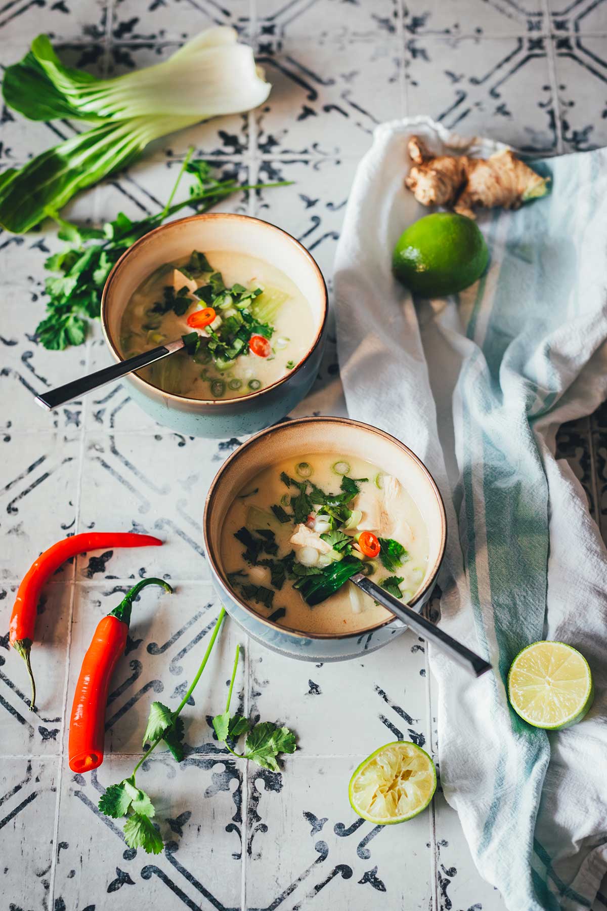 Zwei Schalen Thai-Kokos-Suppe mit Huhn, Pak Choi und Chili, serviert mit Limetten, Ingwer und Koriander auf einem rustikalen Untergrund | moeyskitchen.com