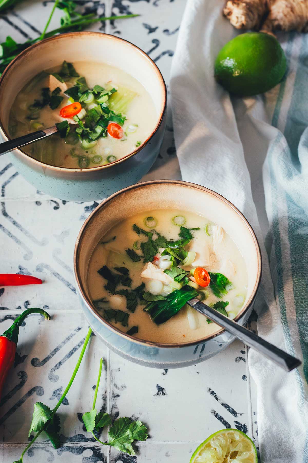 Zwei Schalen Thai-Kokos-Suppe mit Huhn, Pak Choi und Chili, serviert mit Limetten, Ingwer und Koriander auf einem rustikalen Untergrund | moeyskitchen.com