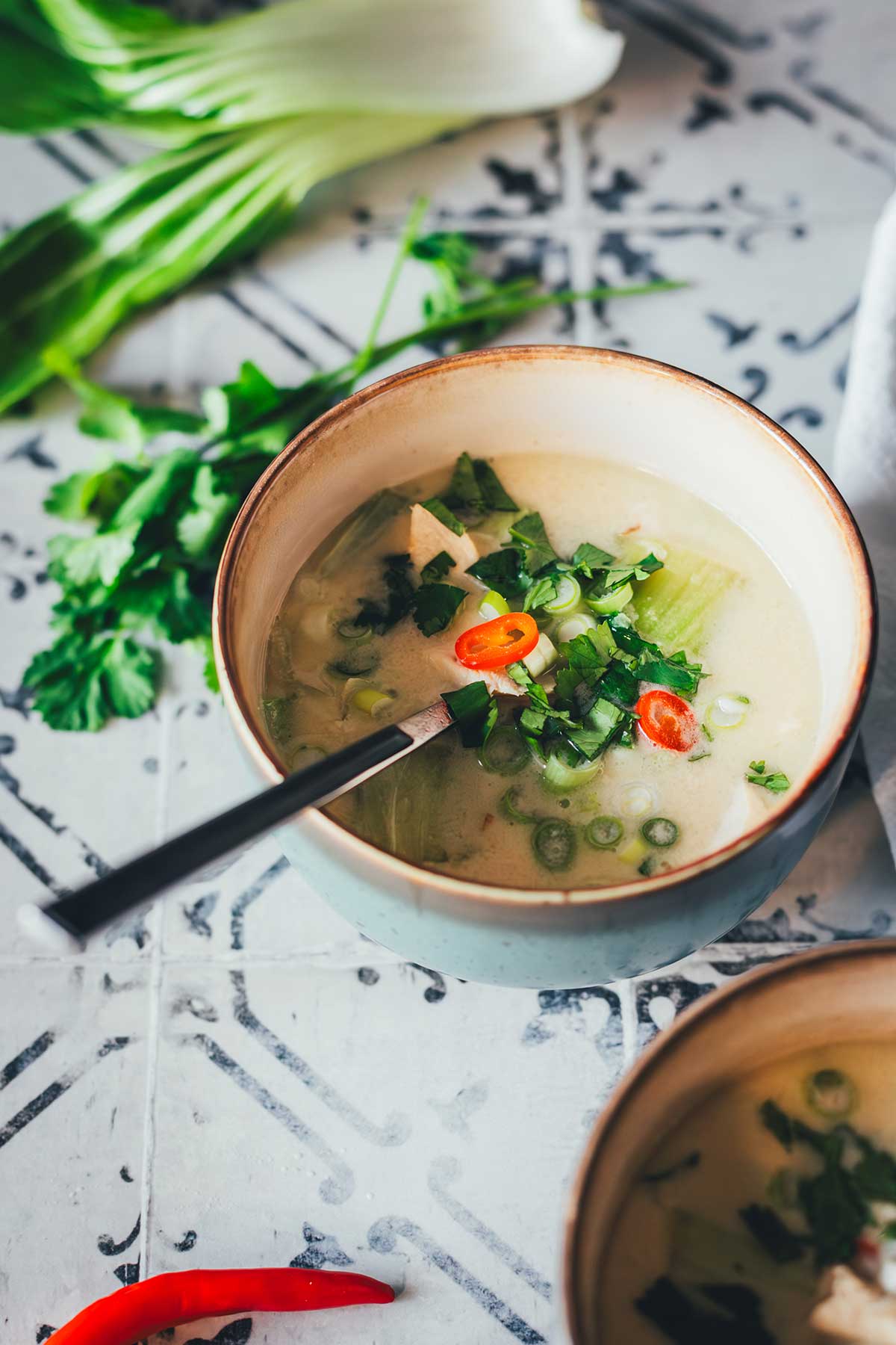 Zwei Schalen Thai-Kokos-Suppe mit Huhn, Pak Choi und Chili, serviert mit Limetten, Ingwer und Koriander auf einem rustikalen Untergrund | moeyskitchen.com