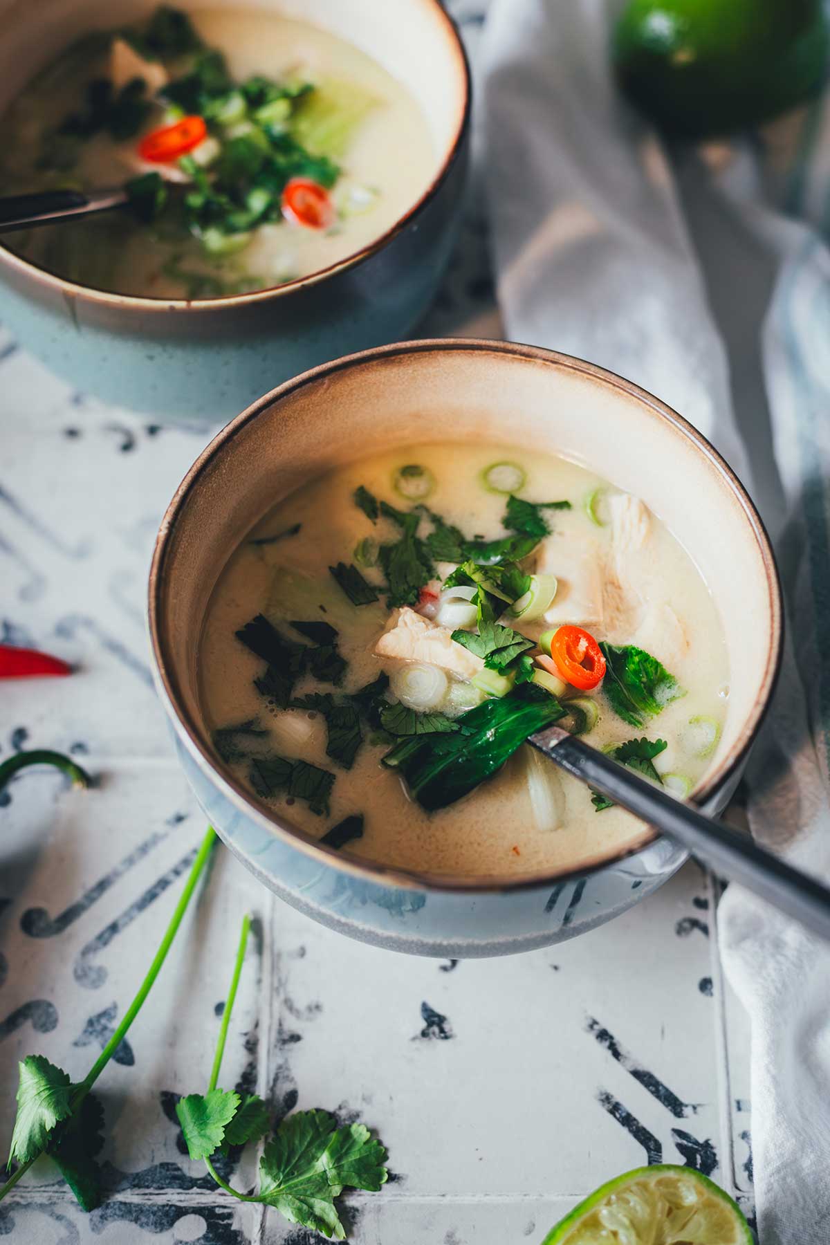 Zwei Schalen Thai-Kokos-Suppe mit Huhn, Pak Choi und Chili, serviert mit Limetten, Ingwer und Koriander auf einem rustikalen Untergrund | moeyskitchen.com