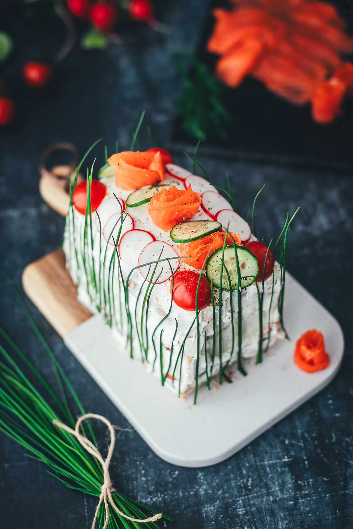 Leichte Draufsicht auf eine Sandwichtorte aus einem Kastenweißbrot. Bestrichen mit Frischkäsecreme und dekoriert mit Räucherlachs-Rosen, Gurke, Tomate, Radieschen und Schnittlauch, serviert auf einem edlen Brettchen