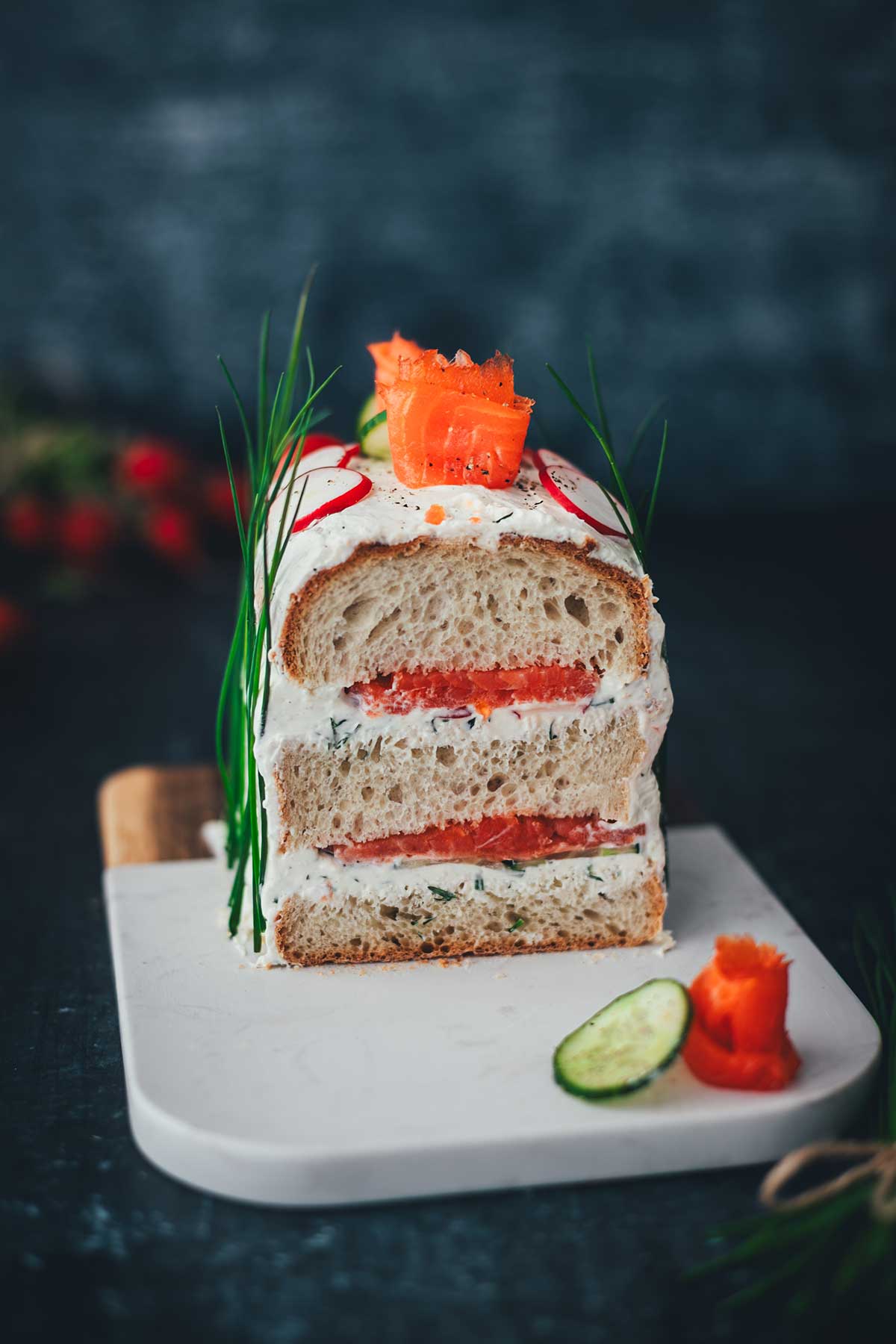 Herzhafte Sandwichtorte nach einer schwedischen Smörgåstårta in der frontalen Ansicht. Man sieht das Kastenweißbrot mit den Schichten aus Frischkäsecreme und Räucherlachs und dazu die Dekoration aus knackigem Gemüse und Schnittlauchhalmen. Angerichtet auf einem hellen Brett vor einem dunklen Hintergrund.