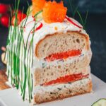 Nahaufnahme einer angeschnittenen Sandwichtorte aus einem Kastenweißbrot, das mit Frischkäsecreme und Räucherlachs gefüllt ist. Die Brottorte macht optisch richtig was her, man sieht die Schichten wie bei einem Kuchen. Dazu ist die mit frischem Gemüse und Schnittlauch dekoriert und auf einem Holzbrett mit Marmor angerichtet.