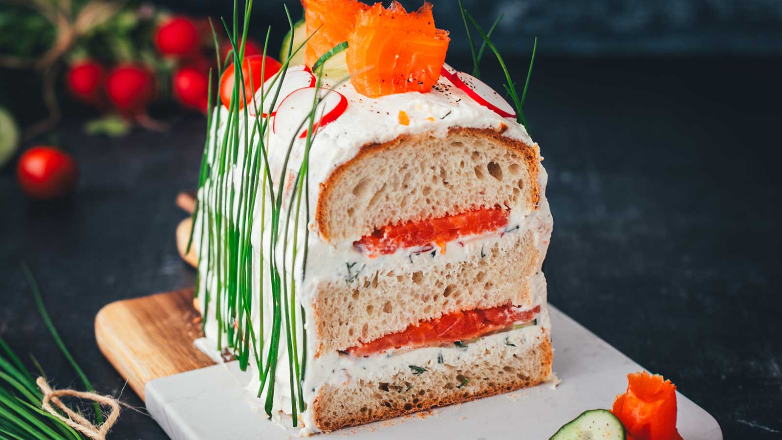 Nahaufnahme einer angeschnittenen Sandwichtorte aus einem Kastenweißbrot, das mit Frischkäsecreme und Räucherlachs gefüllt ist. Die Brottorte macht optisch richtig was her, man sieht die Schichten wie bei einem Kuchen. Dazu ist die mit frischem Gemüse und Schnittlauch dekoriert und auf einem Holzbrett mit Marmor angerichtet.