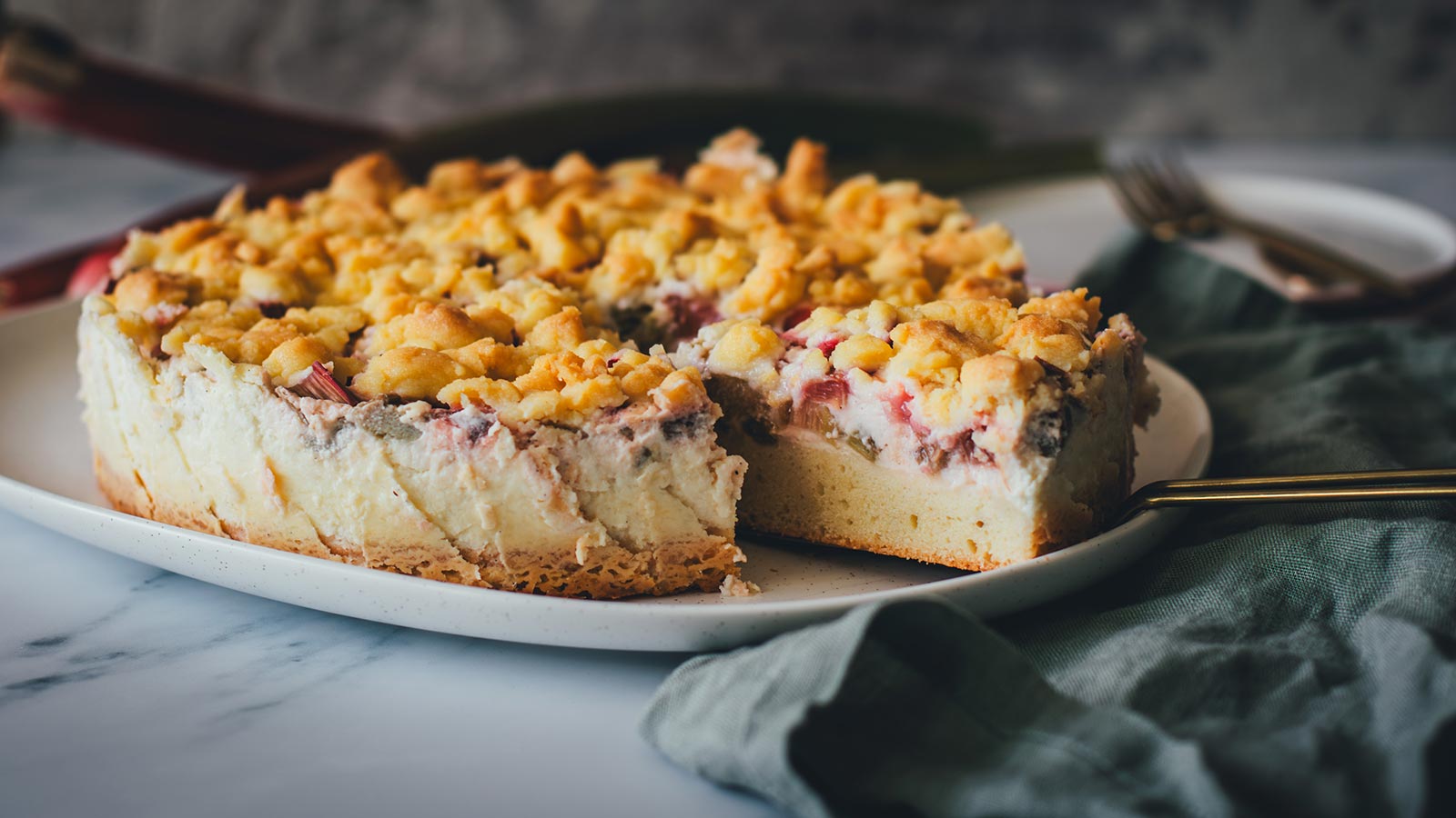 Frisch gebackener Rhabarber-Schmand-Kuchen liegt auf einer Kuchenplatte. Ein Stück wurde herausgeschnitten und liegt servierbereit auf einem Tortenheber. Man sieht den Kuchen im Ganzen und dazu die Schichten aus Rührteig, Schmandcreme, Rhabarber sowie die knusprigen Streusel.