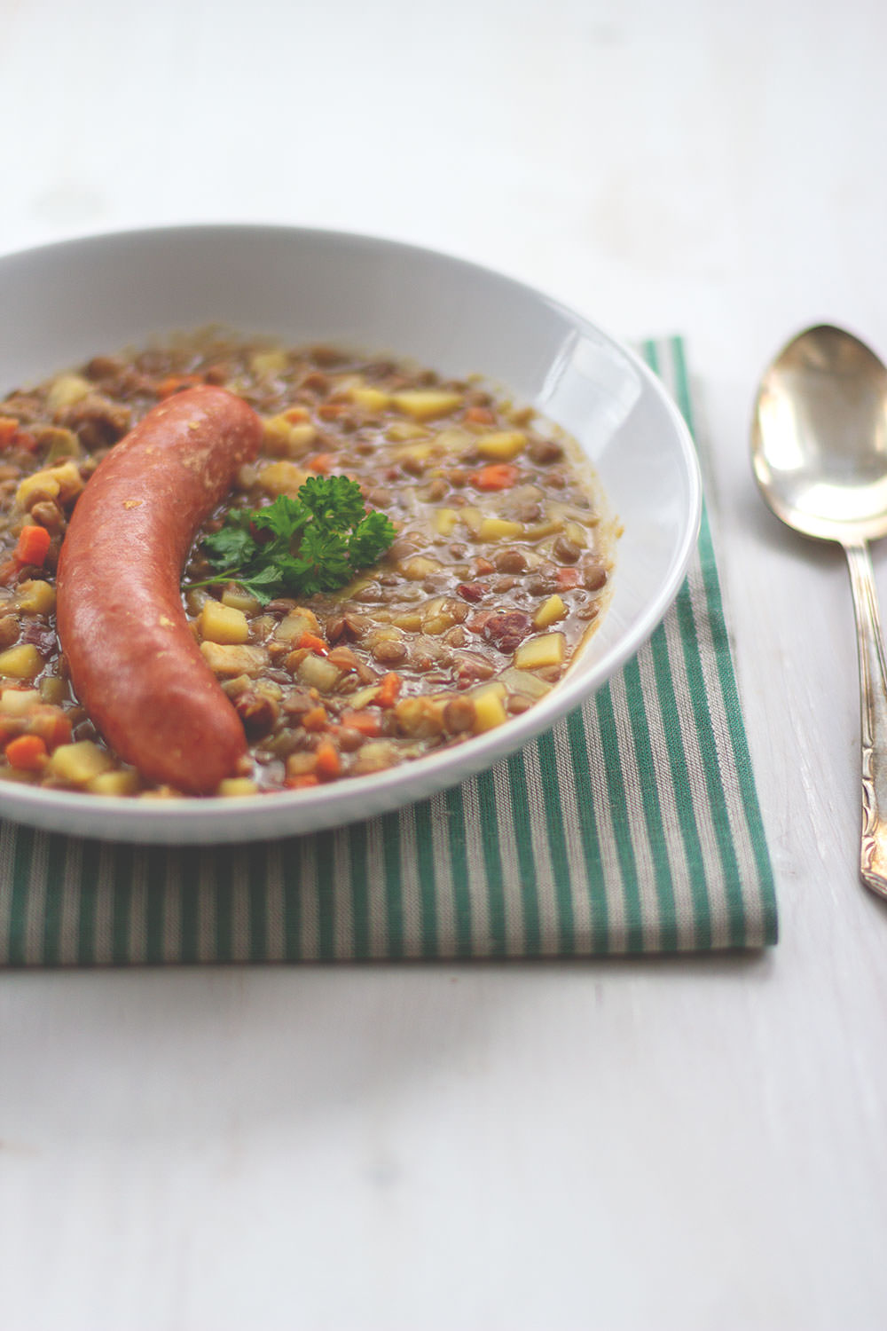Linsensuppe mit Mettenden – der Klassiker, serviert in einem tiefen Teller, bestreut mit etwas frischer Petersilie. Daneben liegt ein Esslöfel. | moeyskitchen.com