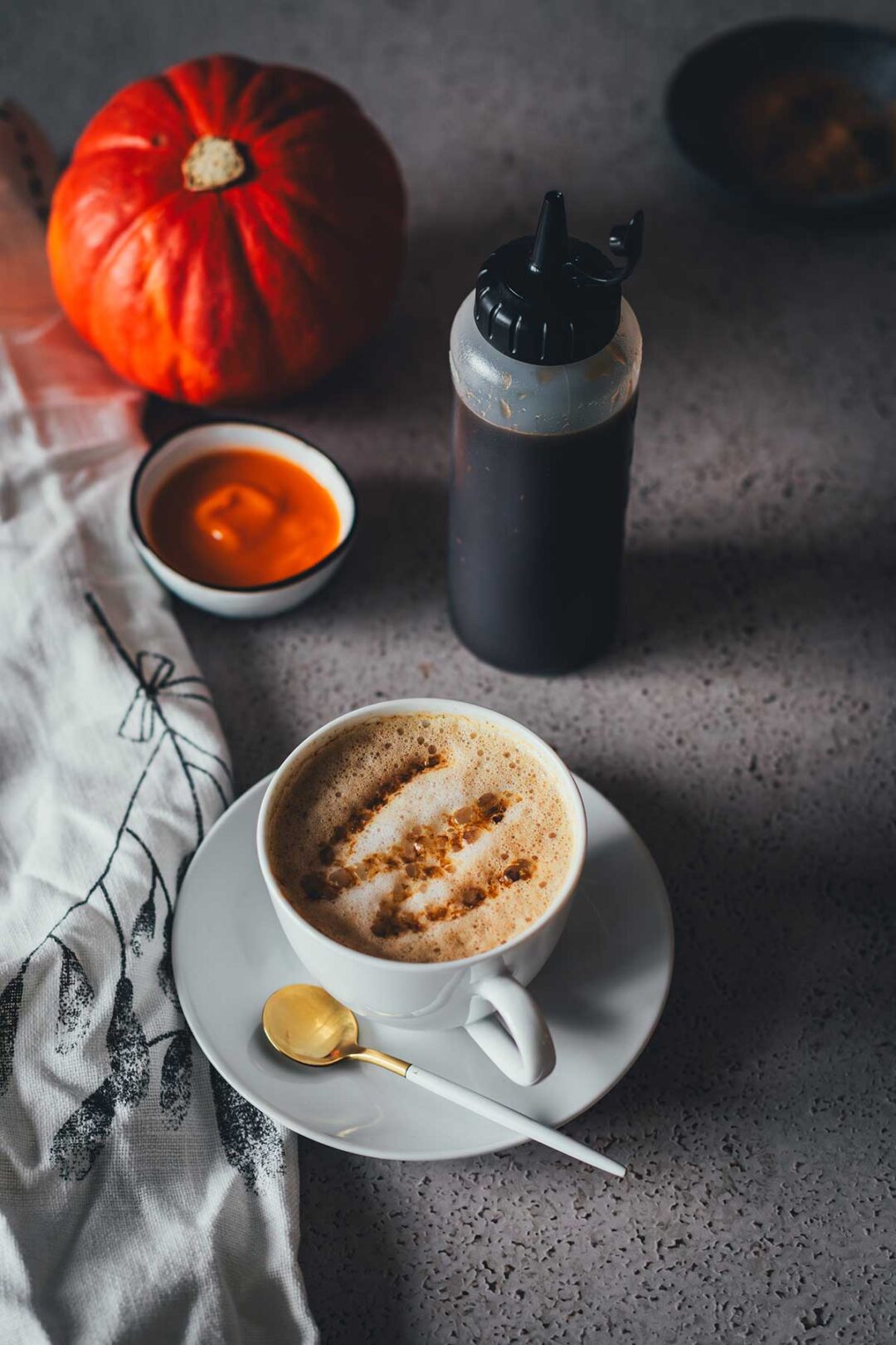 Eine Tasse Cappuccino, getoppt mit Pumpkin Spice Sirup mit brauner Butter, der in eine Squeezeflasche daneben steht. Im Hintergrund sieht man einen Hokkaido Kürbis und Kürbispüree in einer kleinen Schüssel. | moeyskitchen.com