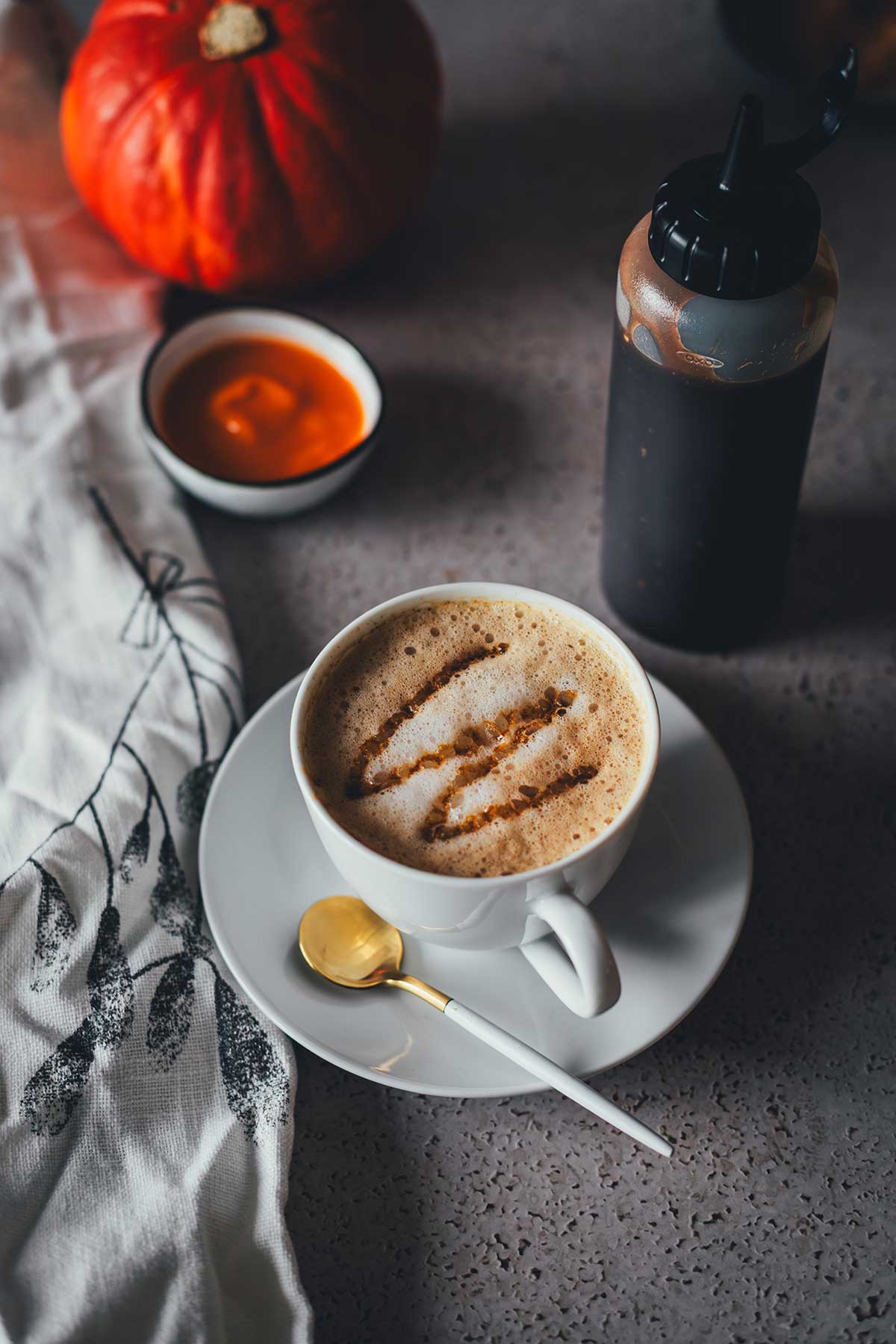 Eine Tasse Kaffee mit Milchschaum, eine Squeezeflasche mit Sirup und eine kleine Schüssel mit Kürbispüree im Hintergrund | moeyskitchen.com