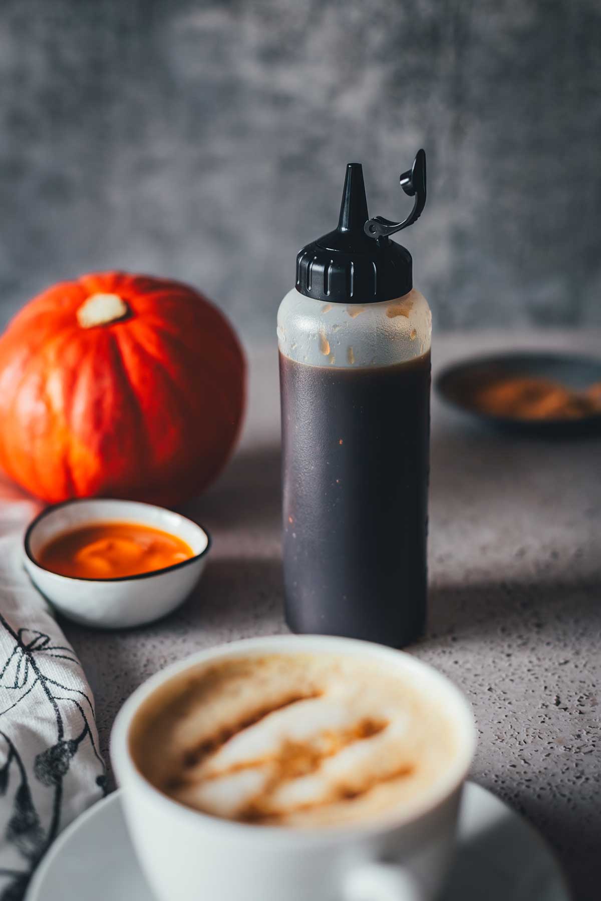 Squeezeflasche mit Pumpkin Spice Sirup im Mittelpunkt. Davor steht eine tasse Cappuccino, im Hintergrund liegt ein Kürbis, eine kleine Schüssel mit Kürbispüree und Gewürze | moeyskitchen.com