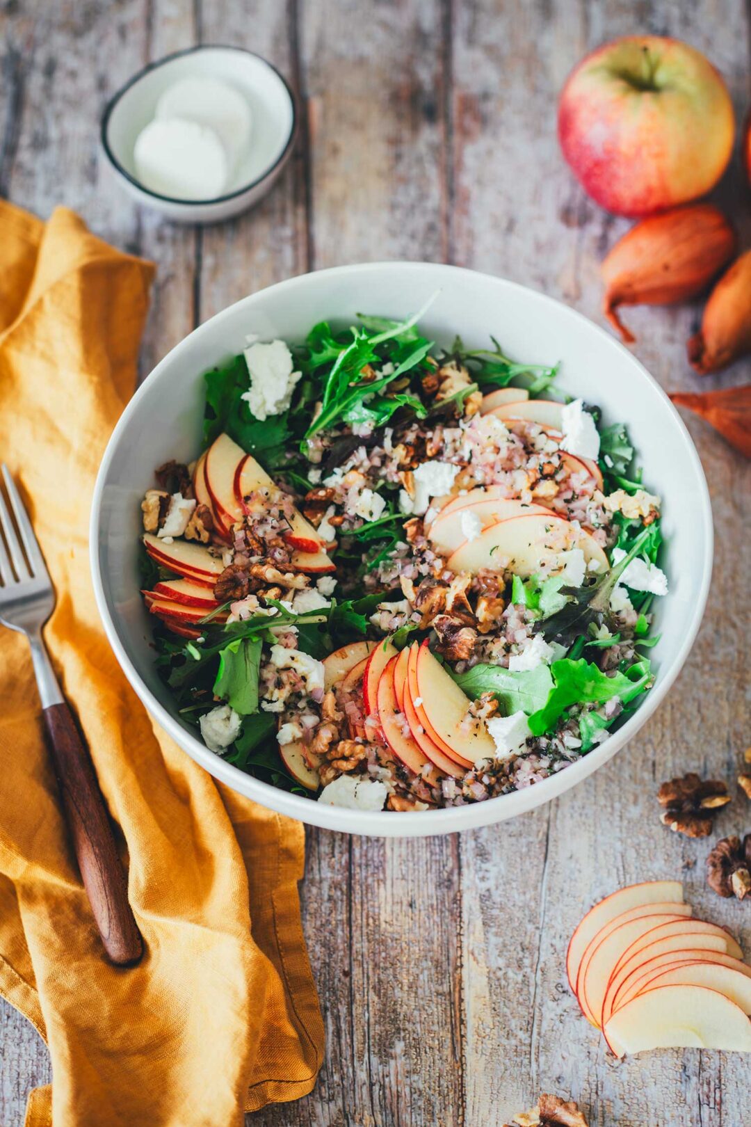 Seitliche Ansicht von einer Salatschüssel mit Herbstsalat – grüner Salat mit Apfel, Ziegenfrischkäse und Walnüssen. Daneben eine gabel mit Holzgriff und eine gelbe Serviette. | moeyskitchen.com