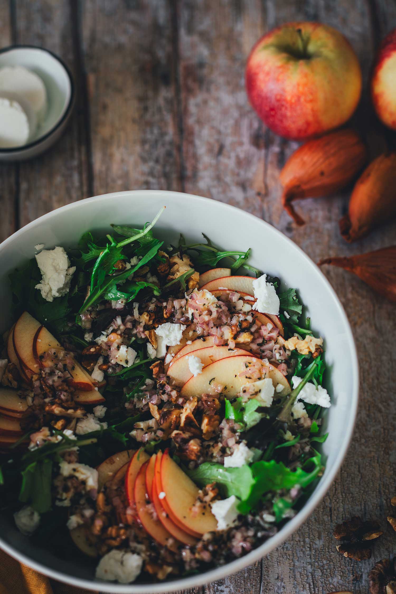 Seitlicher Anschnitt von einer Salatschüssel mit Salat, im Hintergrund Ziegenkäse, Äpfel und Schalotten | moeyskitchen.com