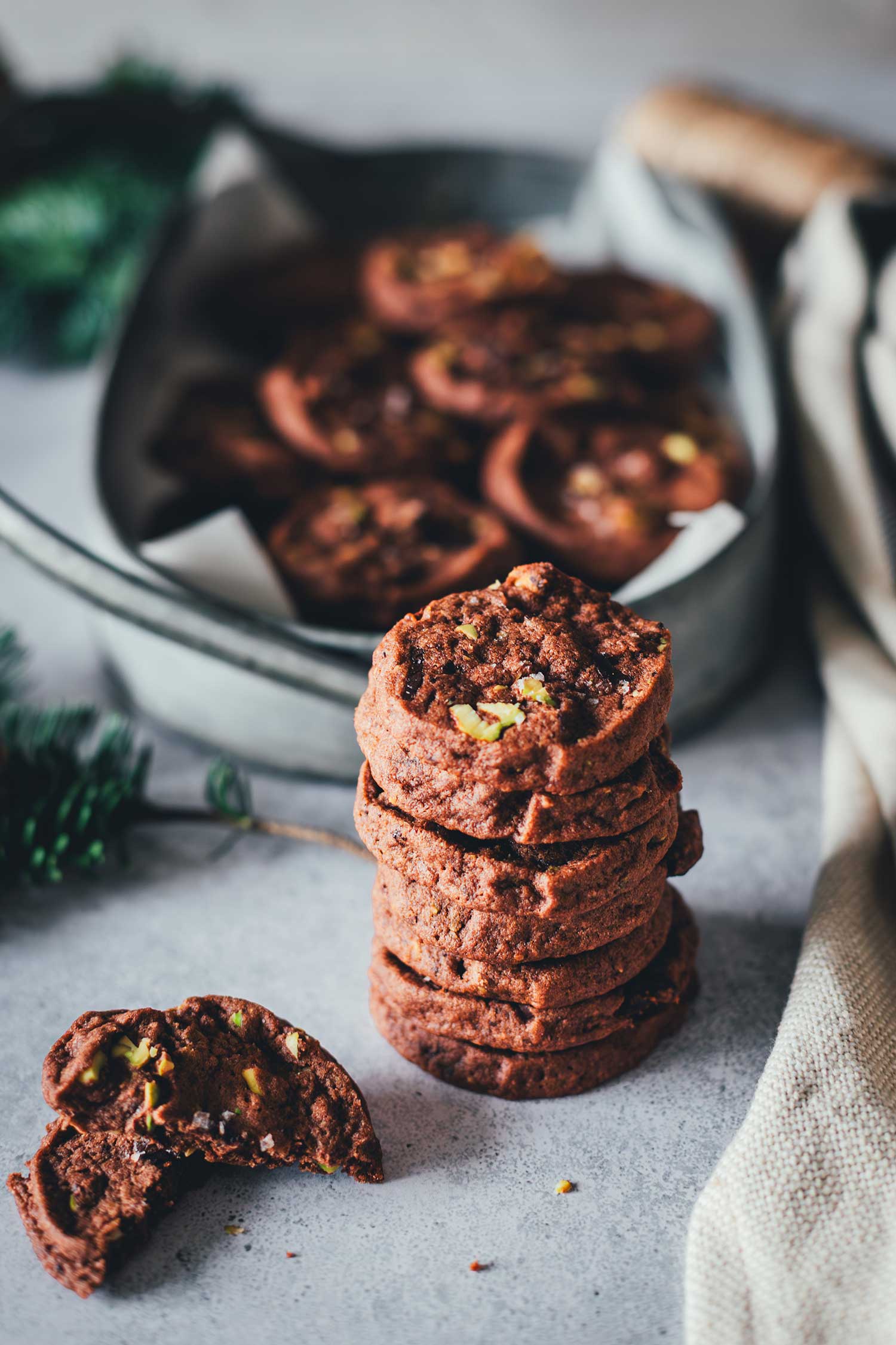 Gestapelte Schokoladen-Pistazien-Cookies auf grauer Oberfläche, daneben ein halb gebrochener Cookie mit sichtbaren Pistazienstückchen; im Hintergrund eine Blechdose mit weiteren Cookies.