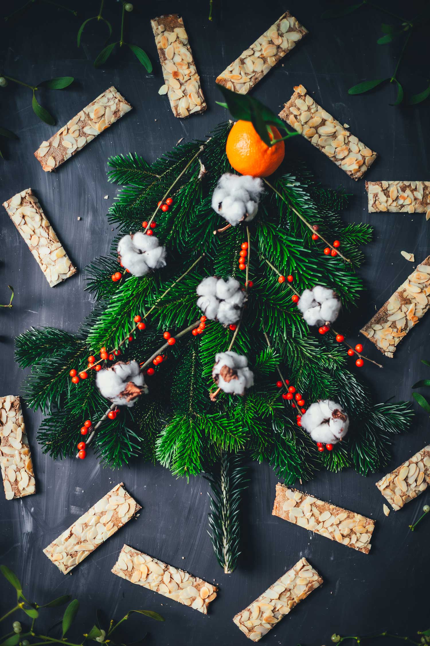 Ein aus Tannenzweigen geformter Weihnachtsbaum liegt auf einem dunklen Untergrund, umgeben von Mandarinen, Deko und Plätzchen | moeyskitchen.com
