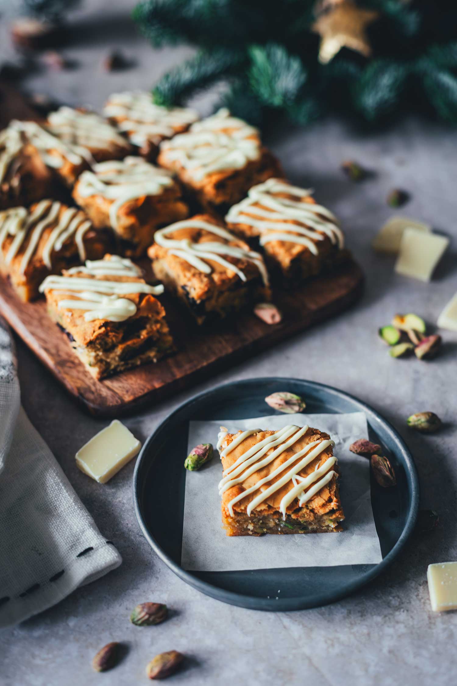 Nahaufnahme von saftigen Weihnachts-Blondies mit Cranberries und Pistazien auf einem Tellerchen und Holzbrett.
