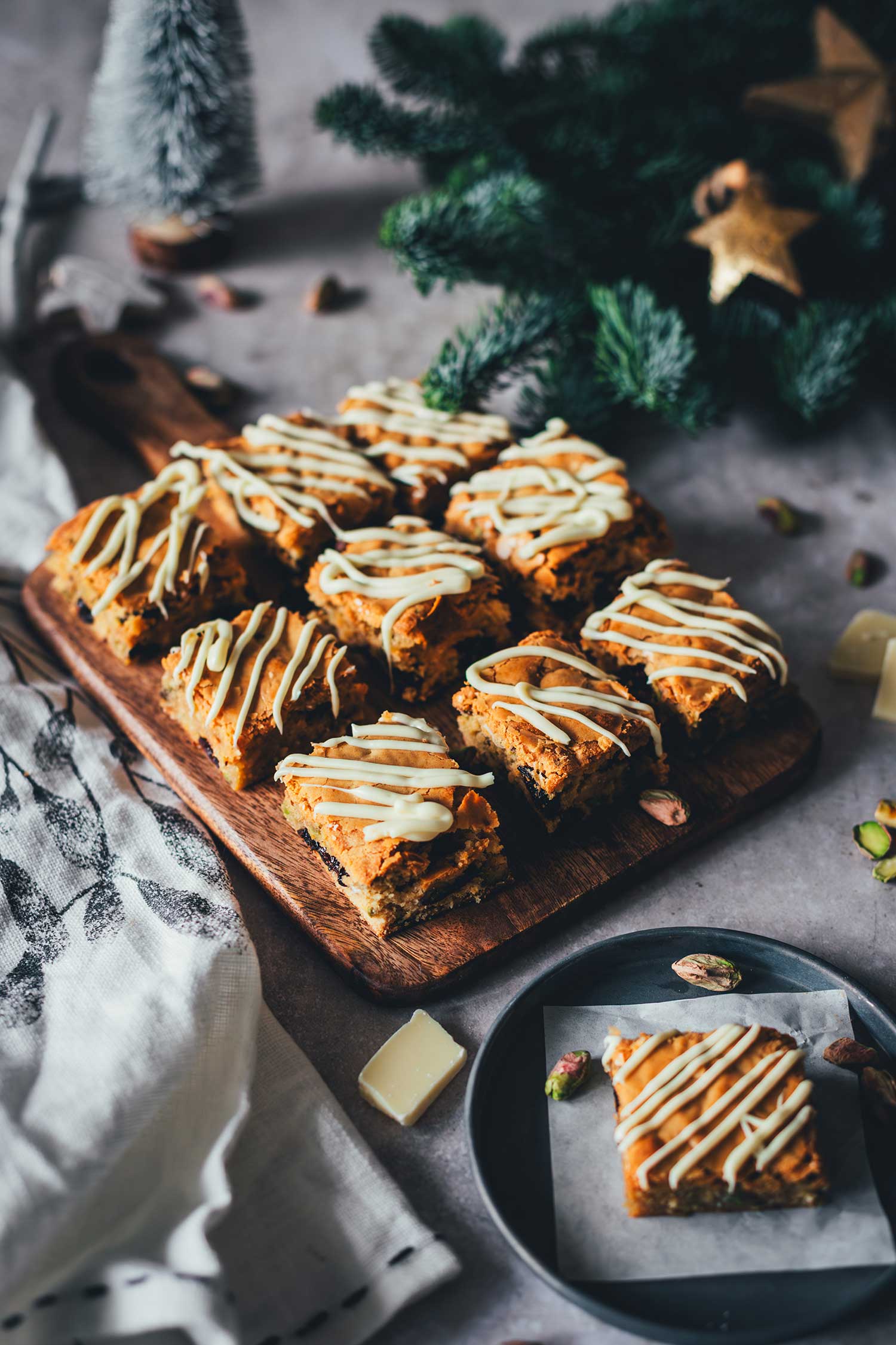 Weihnachts-Blondies auf einem Holzbrett mit einem einzelnen Blondie-Stück auf einem Teller im Vordergrund.