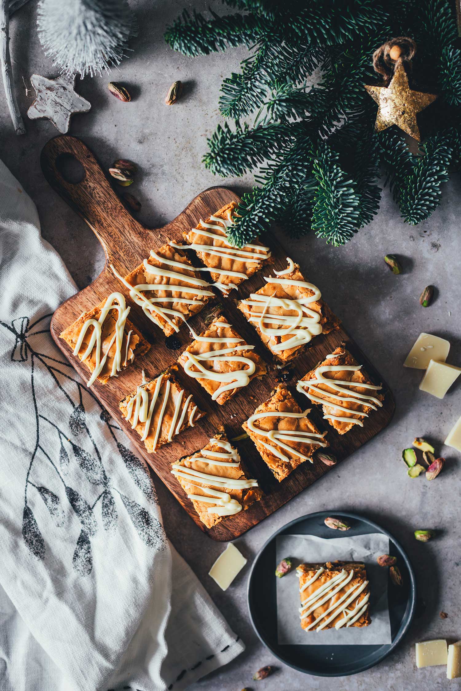 Weihnachts-Blondies mit weißer Schokolade, Cranberries und Pistazien auf einem Holzbrett, von oben fotografiert.