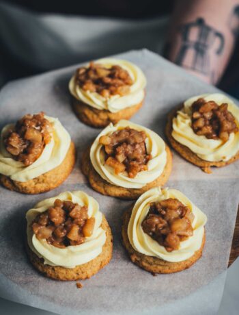 Brown Butter Apple Pie Cookies auf einem Tablett, frisch dekoriert mit Mascarpone-Frosting und Apfeltopping; im Hintergrund unscharfer Arm einer Person beim Servieren.