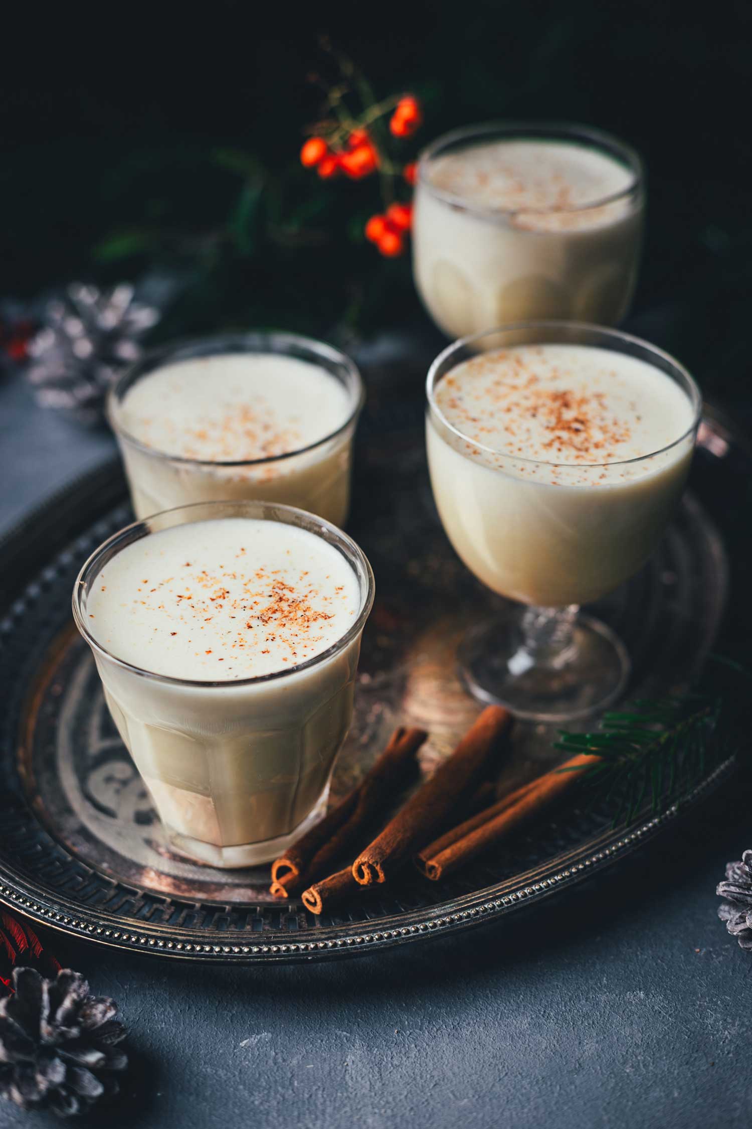Mehrere Gläser mit warmem, cremigem Eggnog, bestreut mit frisch geriebener Muskatnuss, auf einem silbernen Serviertablett angerichtet; daneben Zimtstangen und winterliche Dekoelemente.