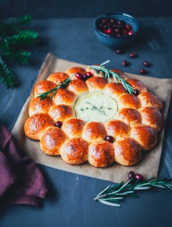 Herzhafter Brioche-Kranz mit geschmolzenem Camembert in der Mitte, dekoriert mit Rosmarin und Cranberries auf dunklem Hintergrund – festlicher Snack für die Adventszeit.