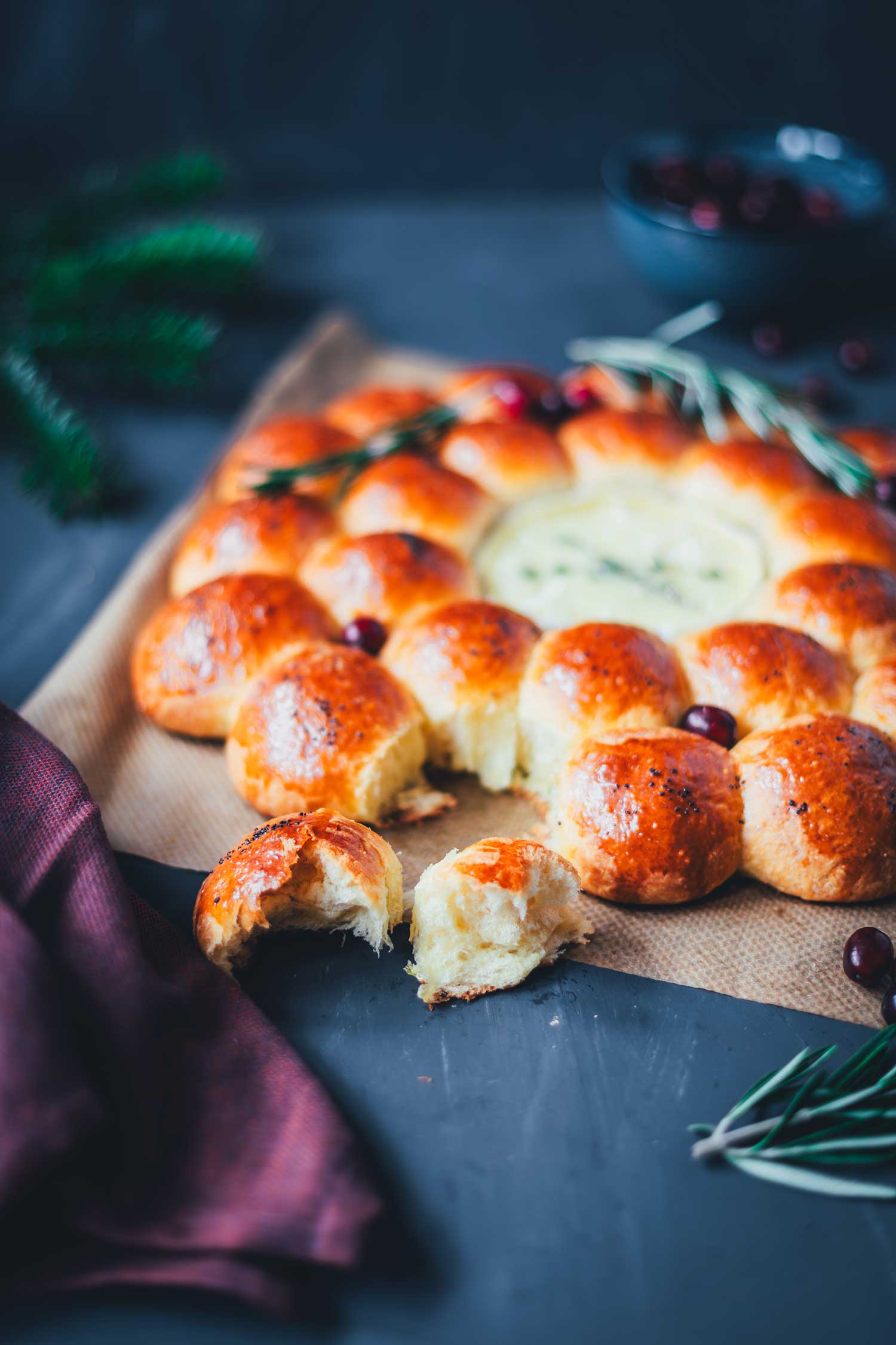 Nahaufnahme eines frisch gebackenen Brioche-Kranzes mit Camembert-Dip; eine abgezupfte, fluffige Teigkugel liegt im Vordergrund, dekoriert mit Rosmarin und Cranberries – festliches Partybrot für die Feiertage.