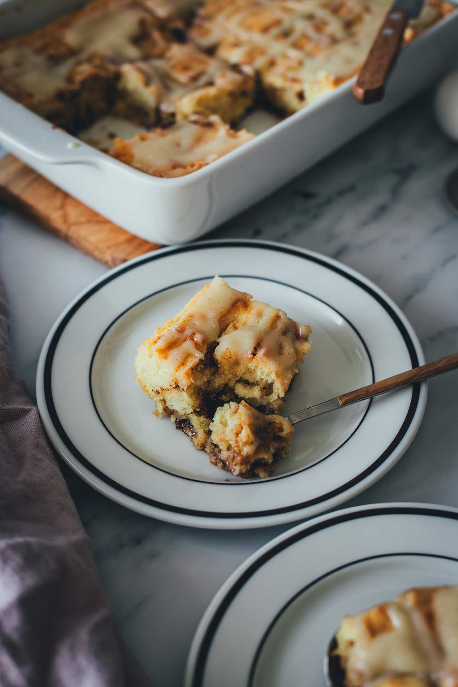 Seitliche Ansicht von einem Stück Zimtschnecken-Kuchen ohne Hefe auf einem Teller. Mit einem Löffel wurde ein Stück abgenommen und man sieht die weiche und saftige Konsistenz des Kuchens. Im Hintergrund steht die Backform.