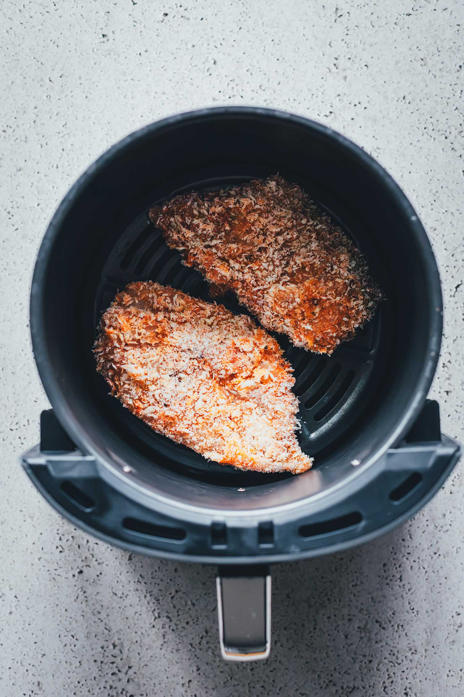 Zwei mit Panko und Paprikapulver panierte Hähnchenbrustfilets im Frittierkorb der Heißluftfritteuse vor dem Garen