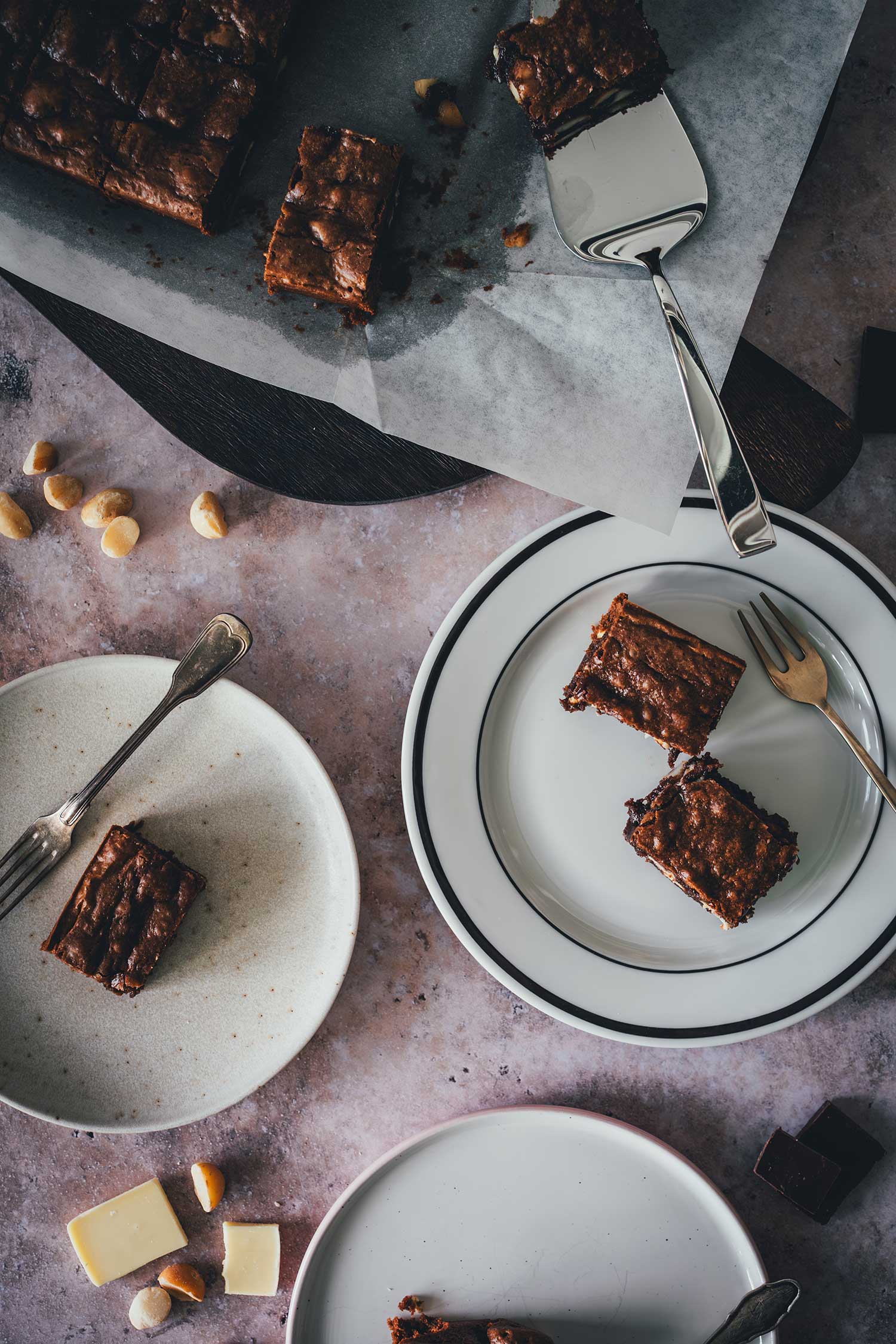 Macadamia Brownies mit weißer Schokolade auf mehreren Tellern angerichtet, mit Tortenheber und verstreuten Macadamias, von oben fotografiert.