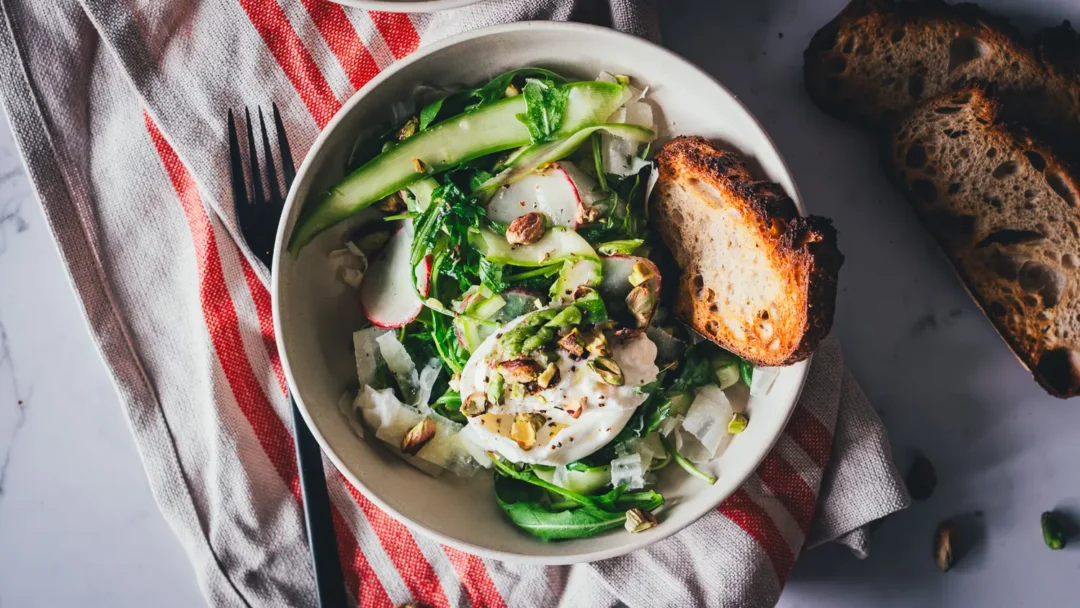 Grüner Spargelsalat mit Burrata, Pistazien und Rucola in einer hellen Schale, daneben getoastetes Sauerteigbrot