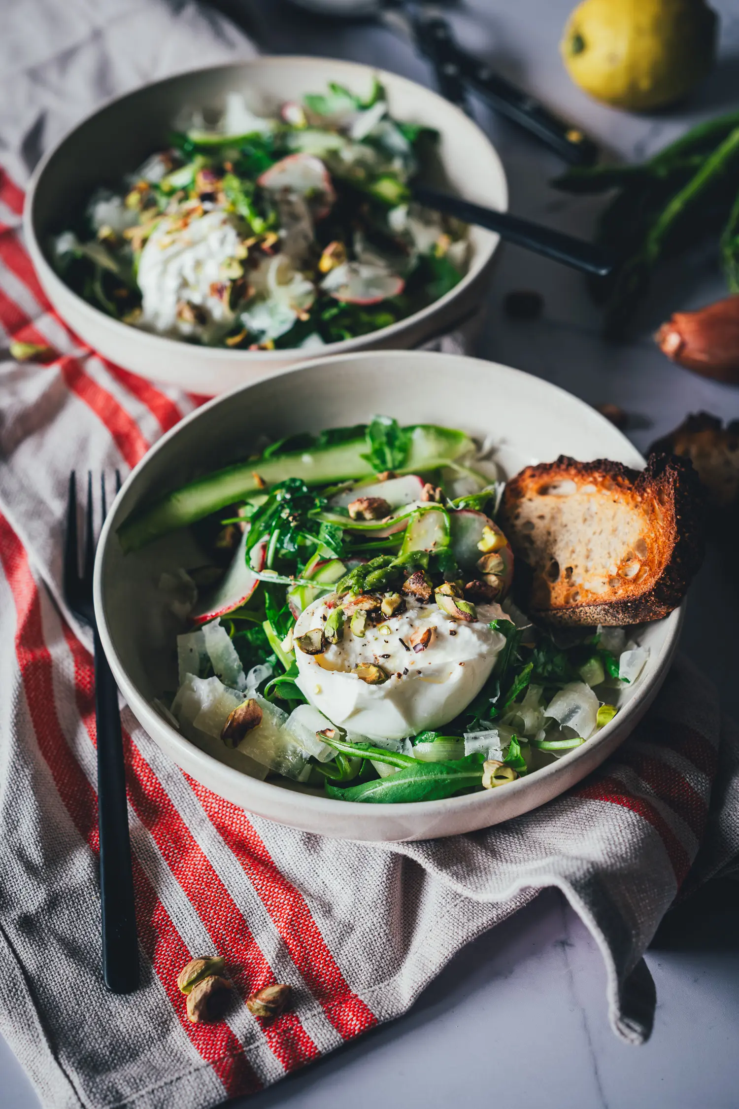 Seitliche Ansicht von zwei frisch angerichteten Tellern mit Spargelsalat aus grünem Spargel, Burrata, Radieschen und Pistazien. Serviert mit etwas geröstetem Brot und auf zwei hellen Tellern serviert.