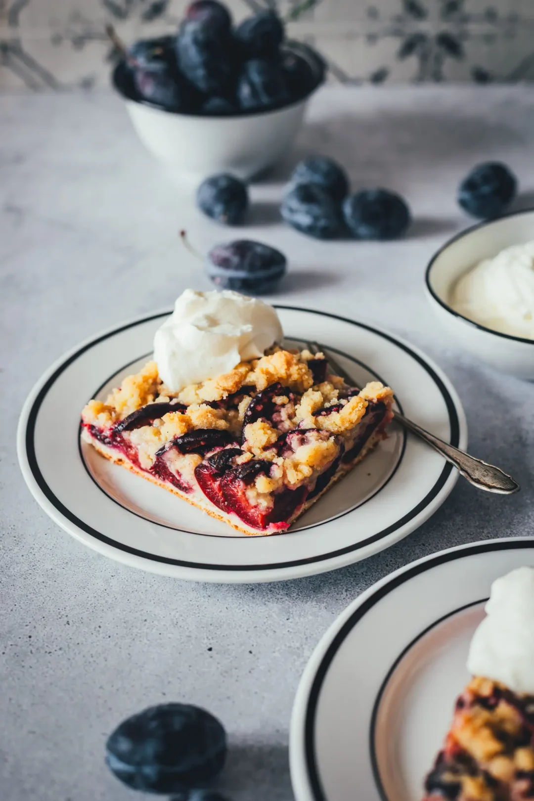 Ein weißer Teller mit einem Stück Zwetschgenkuchen mit Hefeteig und Streuseln, serviert mit Schlagsahne. Im Hintergrund violette Zwetschgen, im Vordergrund ein weiterer Teller.